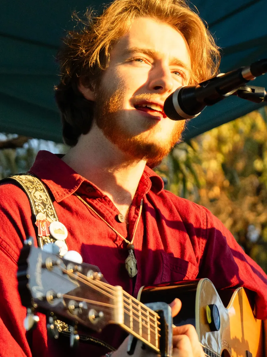 Luke Chandler singing into a microphone with an acoustic guitar, lit by late afternoon sun