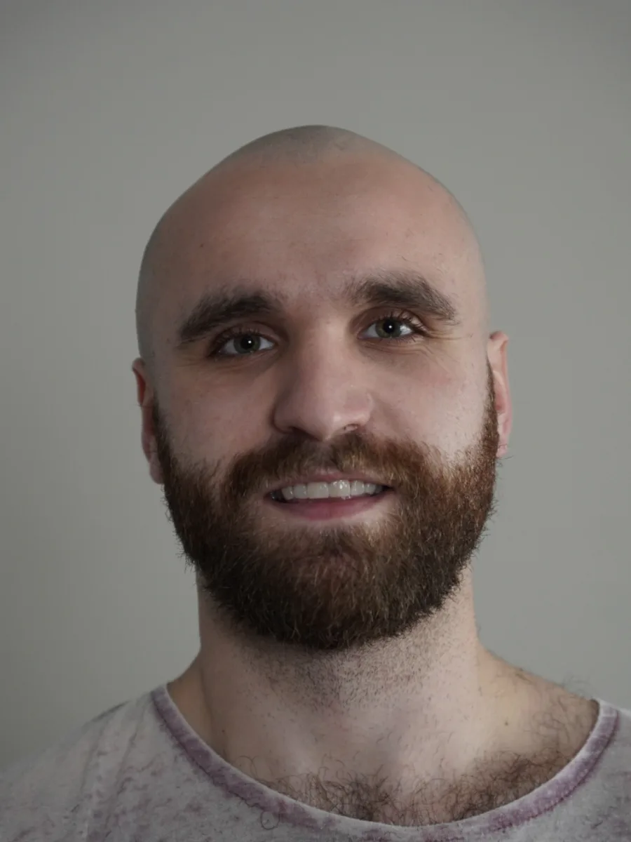 Yannai Goldberg, head shaved and bearded, smiling straight down the lens in front of a plain grey wall