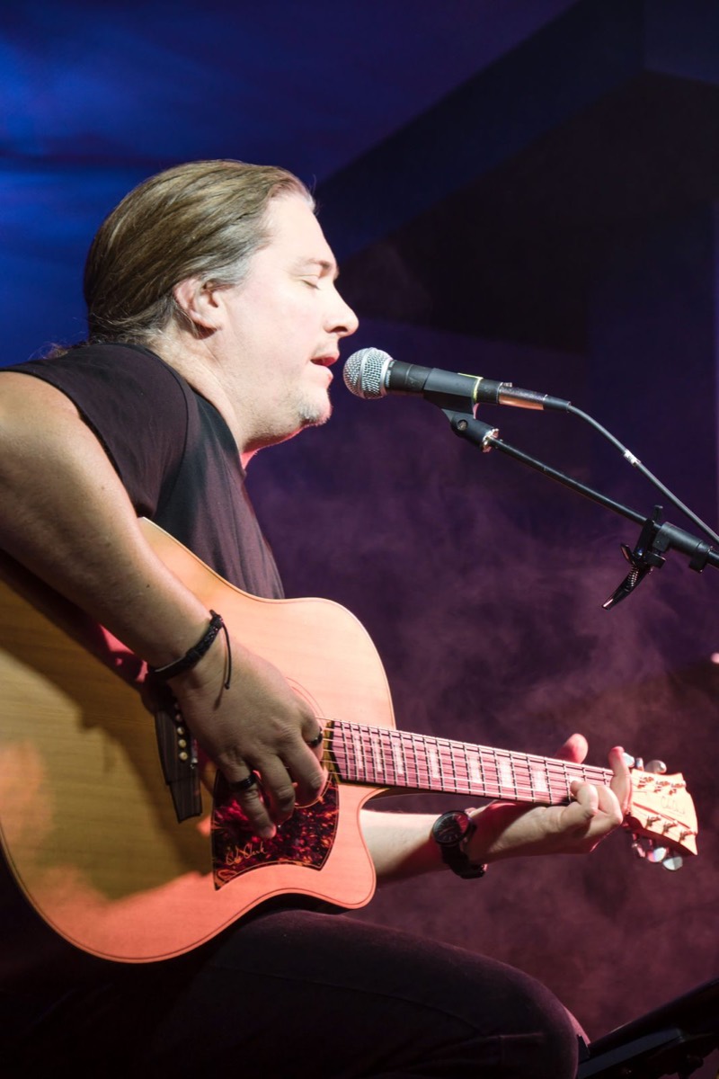 Seated guitarist singing into the mic through stage haze